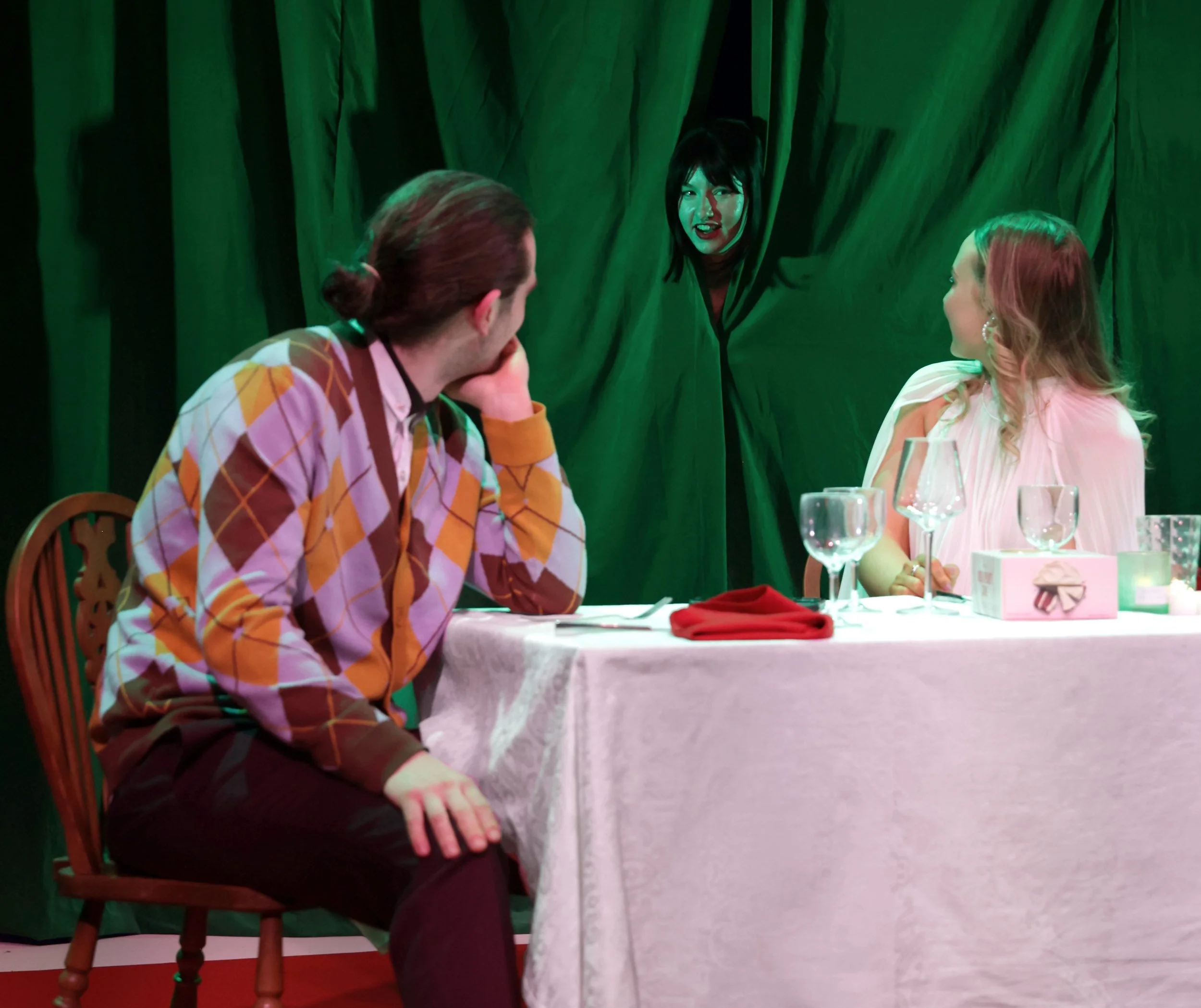 Three actors at a dining table with green curtains in the background. A woman with black hair is looking out from behind the curtain, smiling. The other two - a man and a woman - are looking back at her.