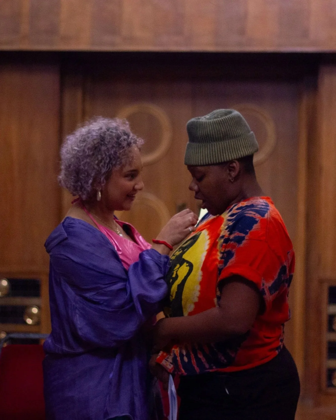Two Black actors are facing each other , one nonbinary person with curly lavender hair in a purple jacket and pink top, the other a woman with a beanie hat and a colorful tie-dye shirt. They are sharing a soft, romantic moment