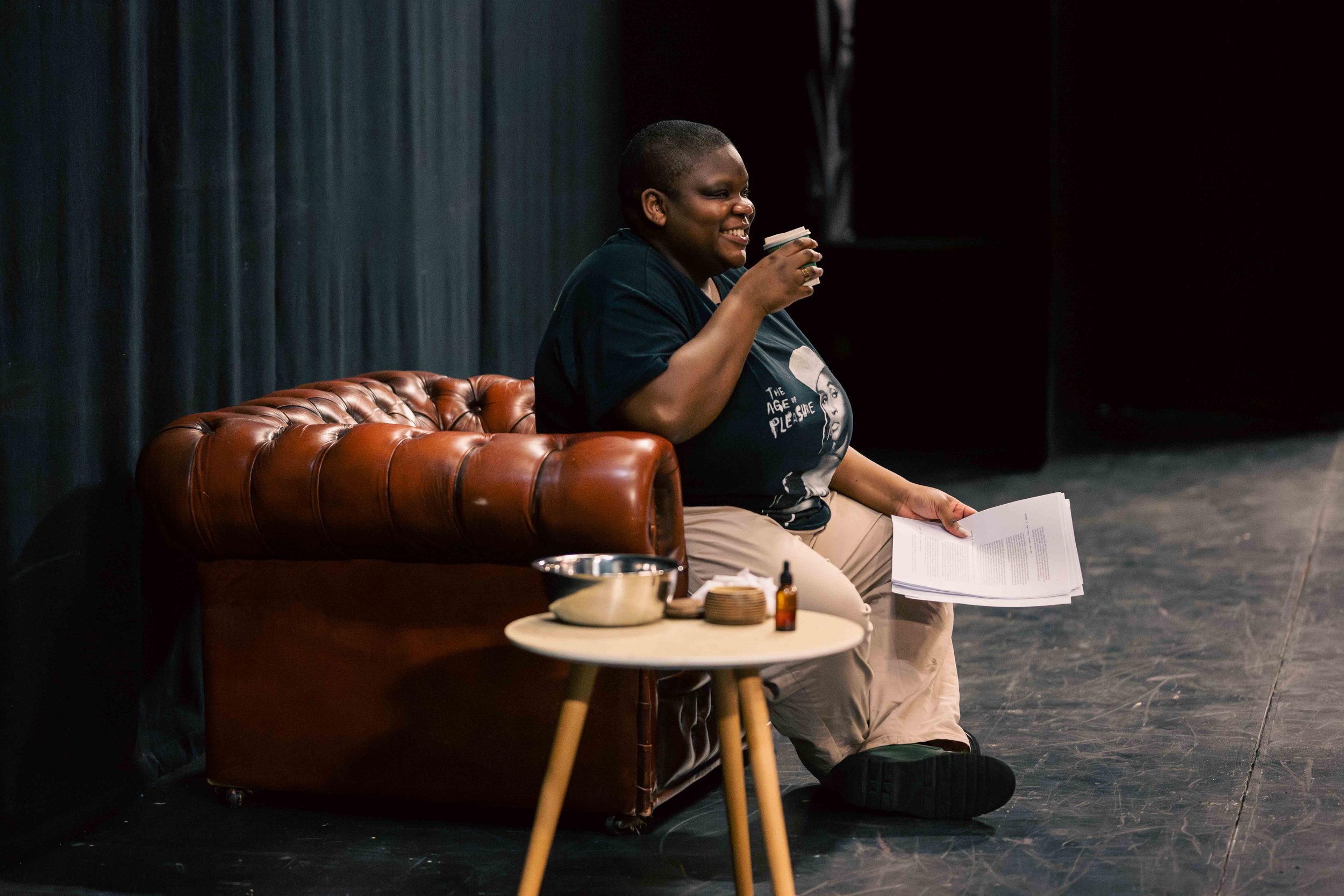 Maz - a fat Black nonbinary person - sitting on a brown leather couch, smiling, holding a coffee cup, holding a script, on stage with a black curtain backdrop.