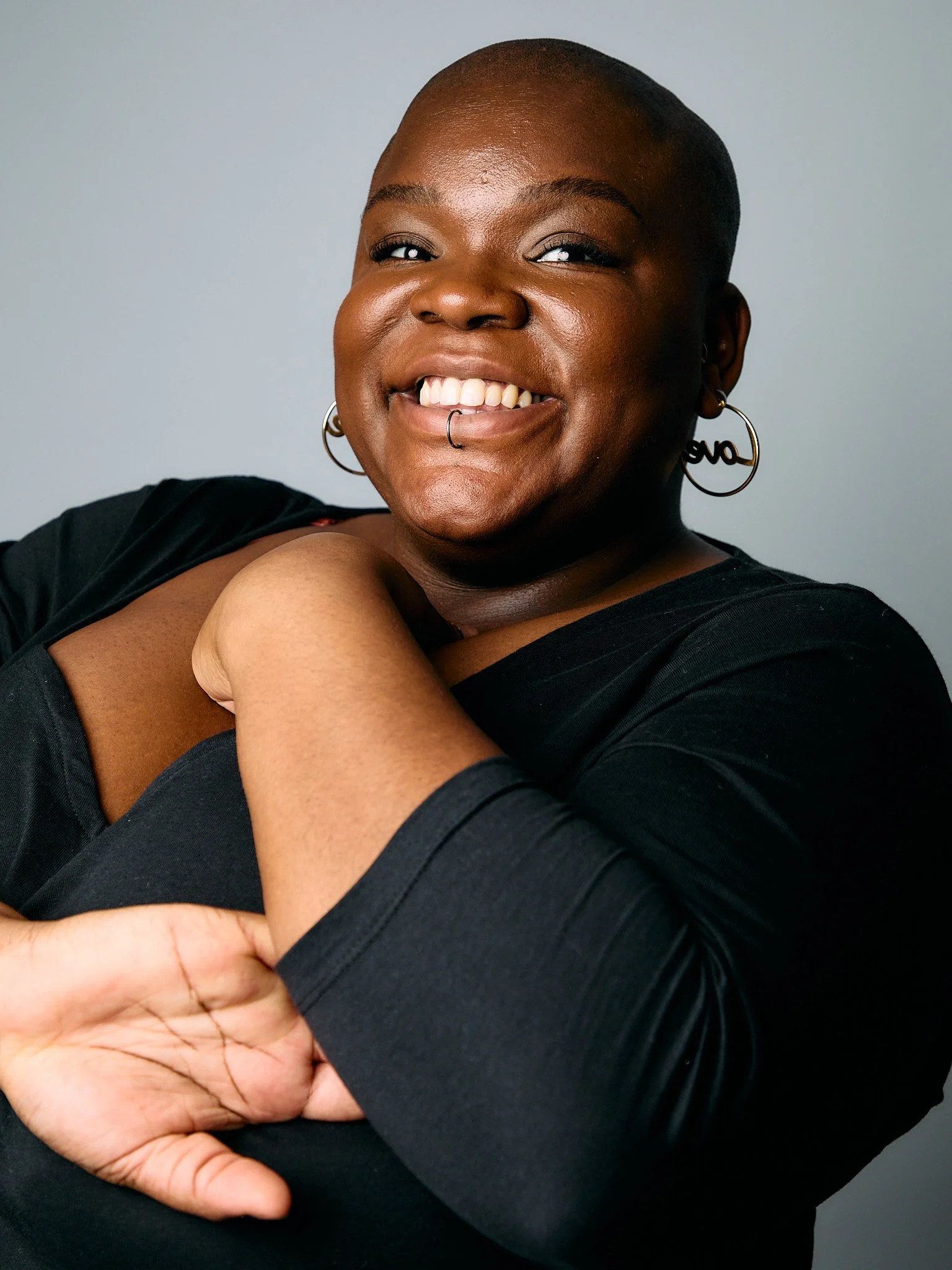 Maz - a fat Black nonbinary person with a shaved head and large hoop earrings -  wearing a black top, smiling against a plain gray background.