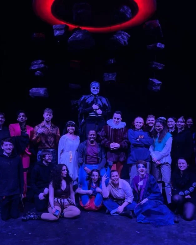 Group of people on stage with a person dressed as a clown in the center. The background is dark with hanging rocks and a red circular installation above.