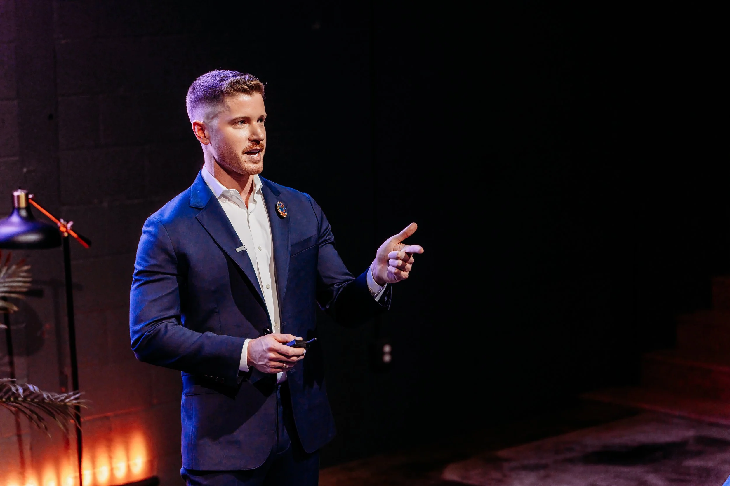 Blake Mitchell in a suit speaking and gesturing on a dimly lit stage