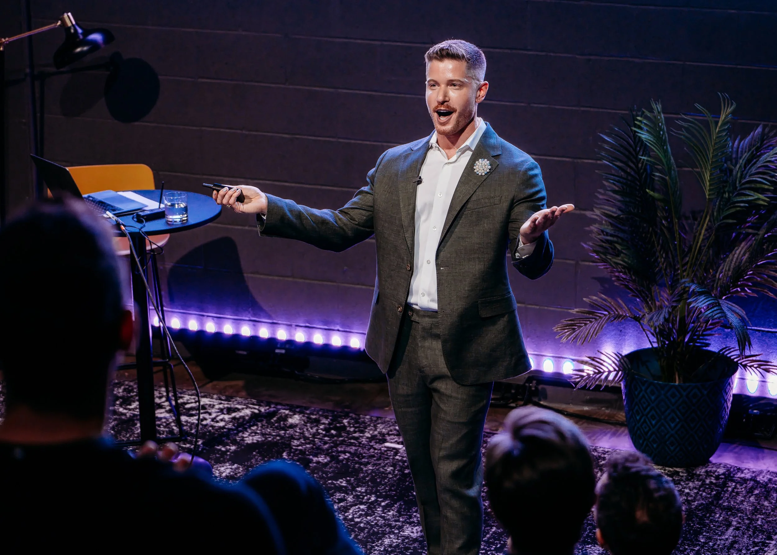 Blake Mitchell speaking to an audience on a dimly lit stage, holding a presentation clicker beside a table and potted palm.