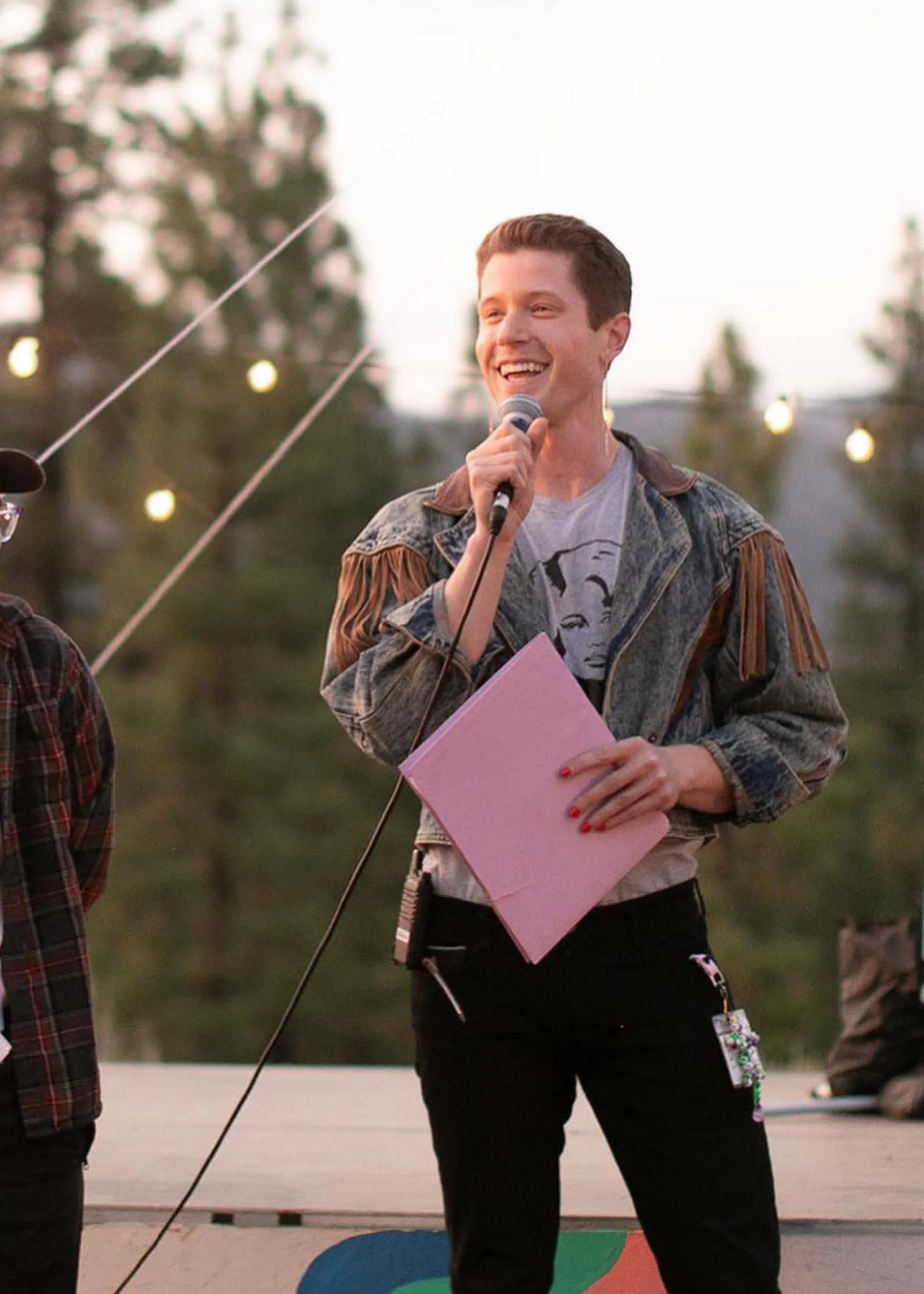 Blake Mitchell holding a microphone and pink folder at an outdoor event with string lights