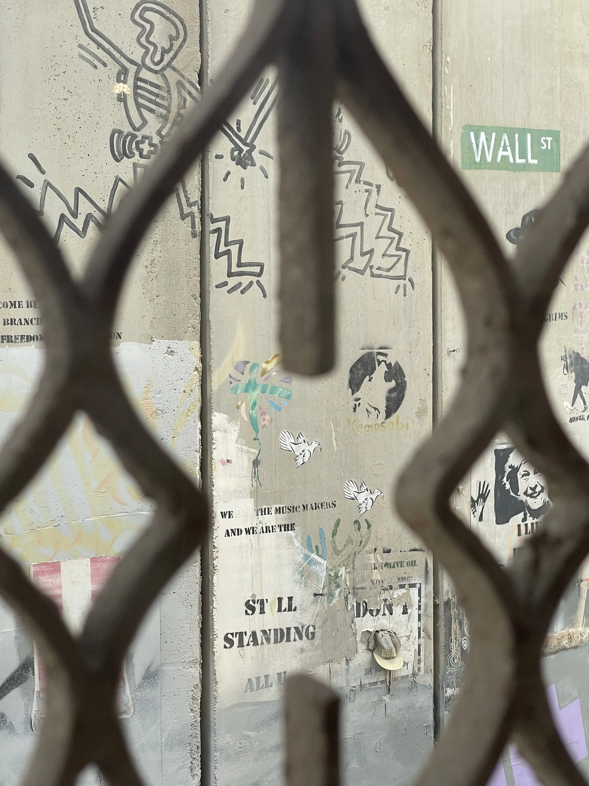 Graffiti-covered concrete wall seen through a chain-link fence, featuring a green Wall Street sign and layered posters.