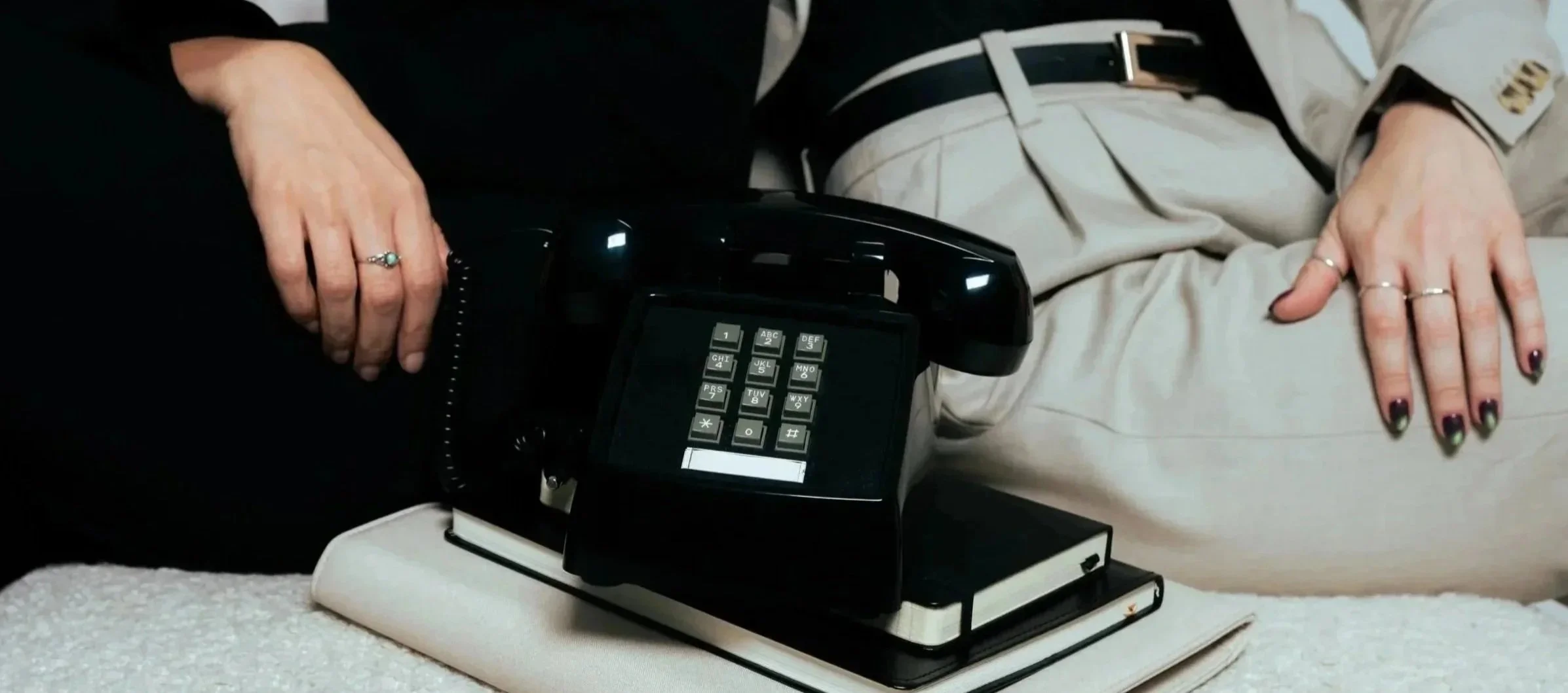 A person dressed in light-colored pants and ringed fingers resting on their lap next to a black rotary desk phone on top of a closed notebook.