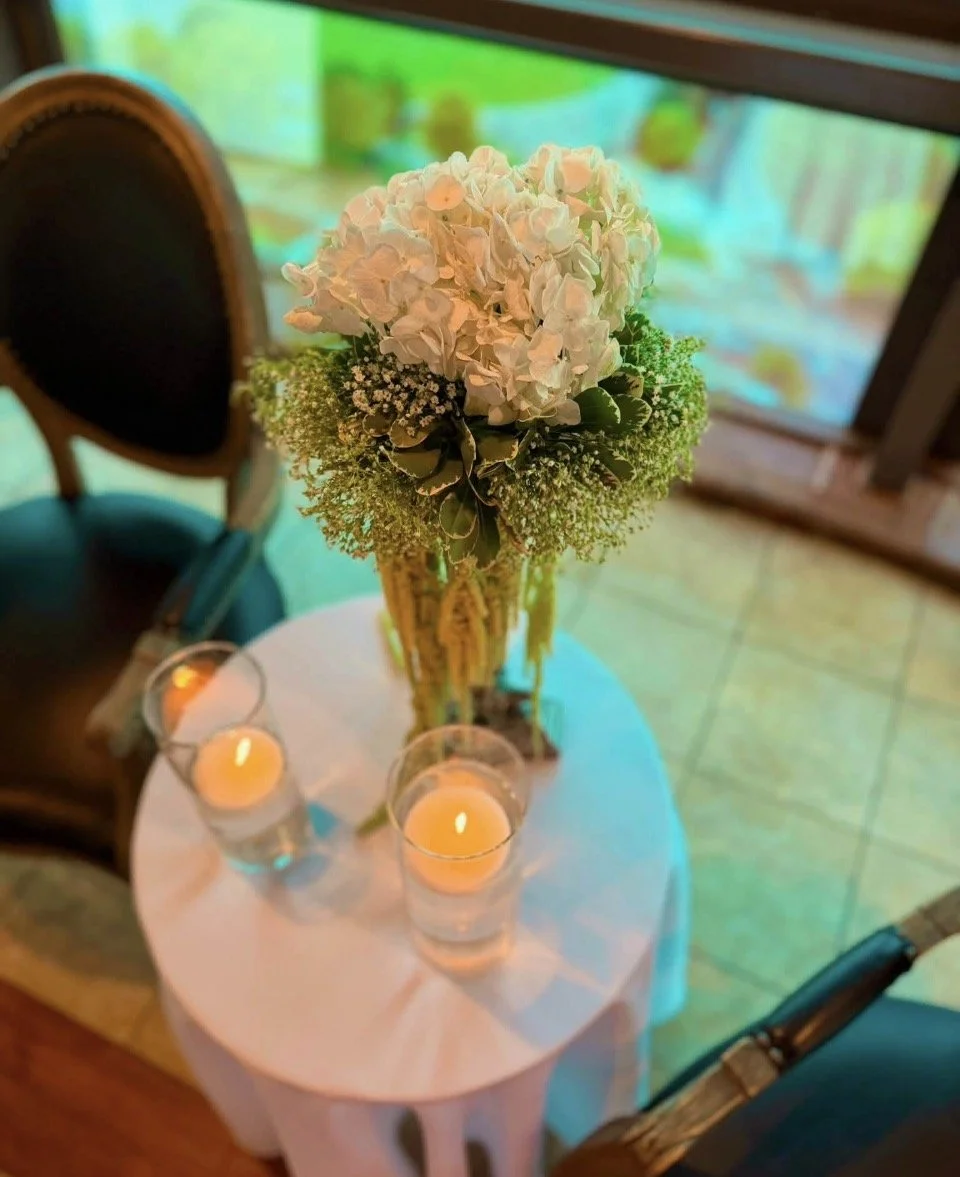 A floral arrangement of white hydrangeas and baby's breath on a round table with a white tablecloth, accompanied by three lit candles in glass holders. In the background, there is a window or glass door, with a green-tinted outdoor scene visible.