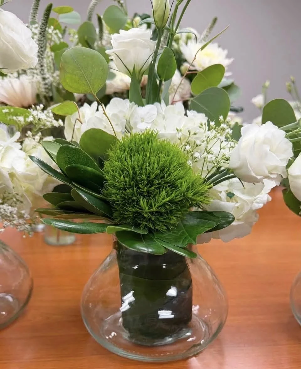 A floral arrangement with white and green flowers and foliage in a glass vase on a wooden surface.