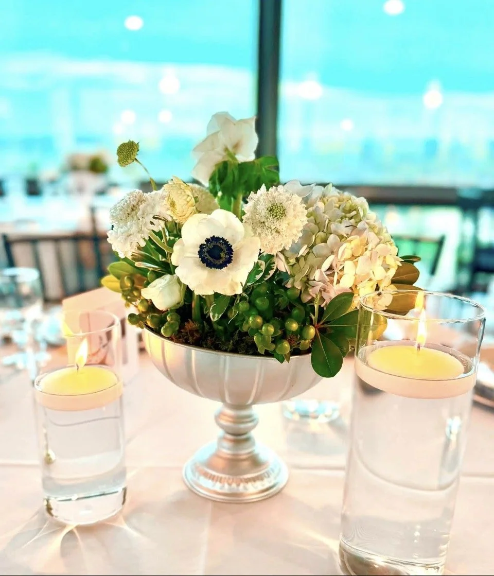 A floral centerpiece with white flowers in a silver vase, surrounded by two floating candles in glass holders, on a table with a light-colored tablecloth.