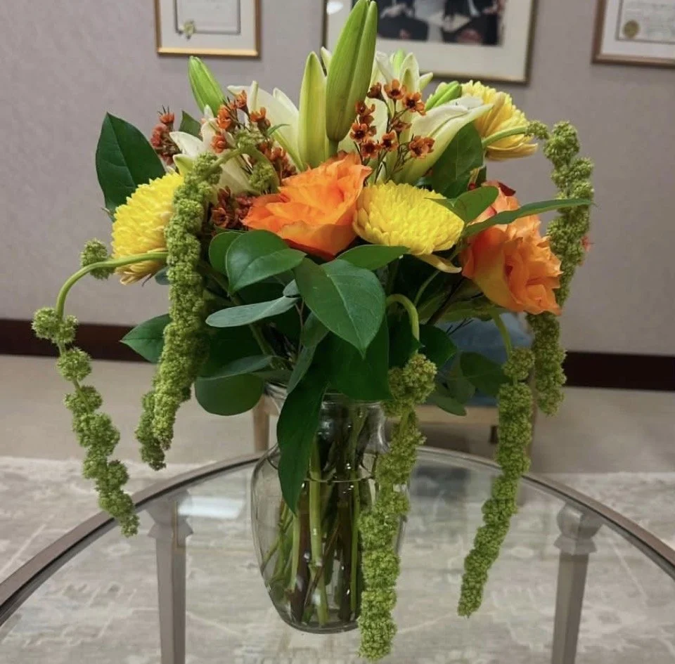 A glass vase filled with a colorful bouquet of flowers, including yellow, orange, and white blooms, with green foliage, placed on a glass table indoors.