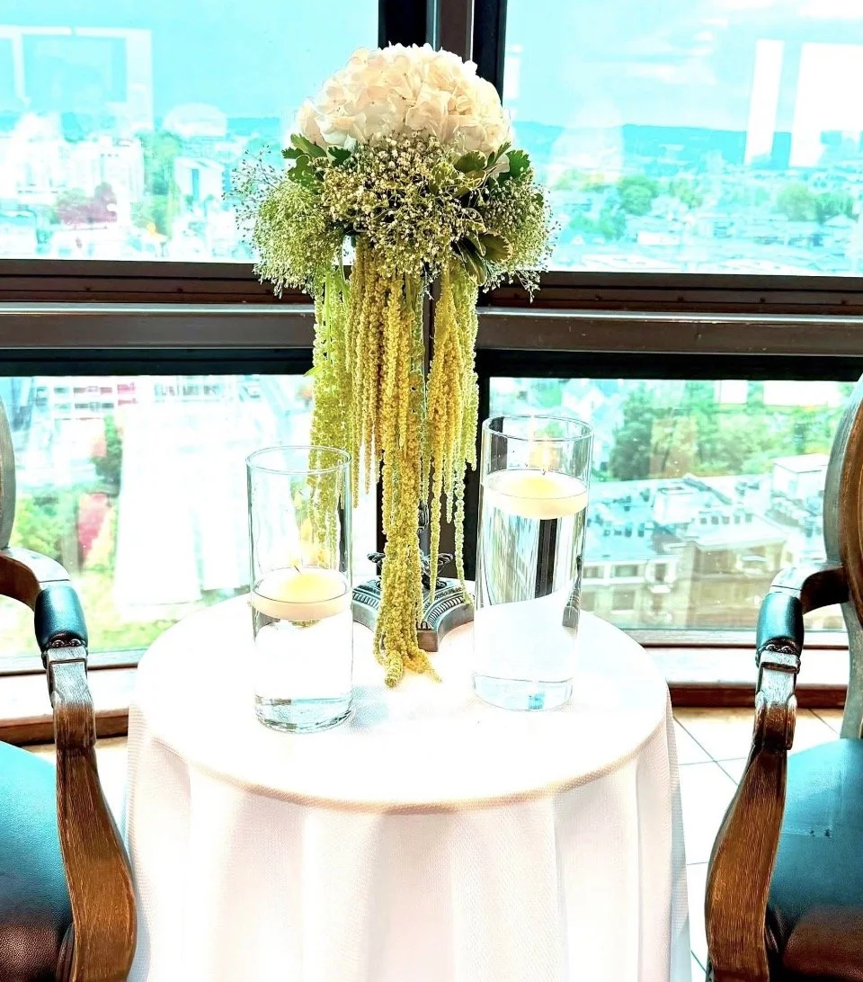A round table with a white tablecloth features a floral centerpiece with white hydrangeas, baby's breath, and hanging yellow flowers. Two glasses of water with floating candles are on the table. Large windows with a city view provide natural light.