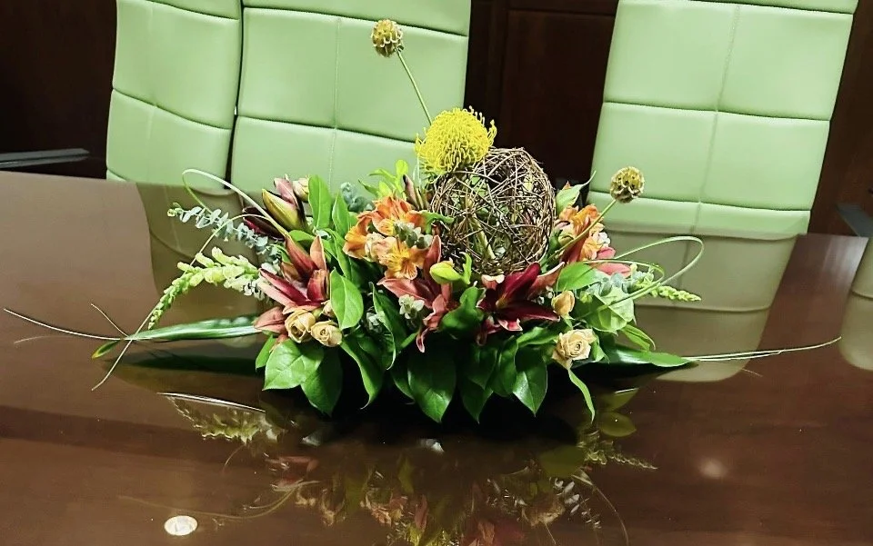 A floral arrangement with yellow, orange, and cream-colored flowers, green leaves, and decorative branches on a polished wooden table in front of green upholstered chairs.