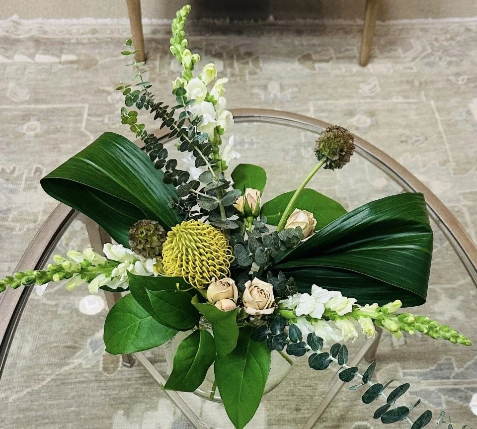 A floral arrangement with large green leaves, white flowers, and small beige roses in a clear glass vase on a round glass-top table.