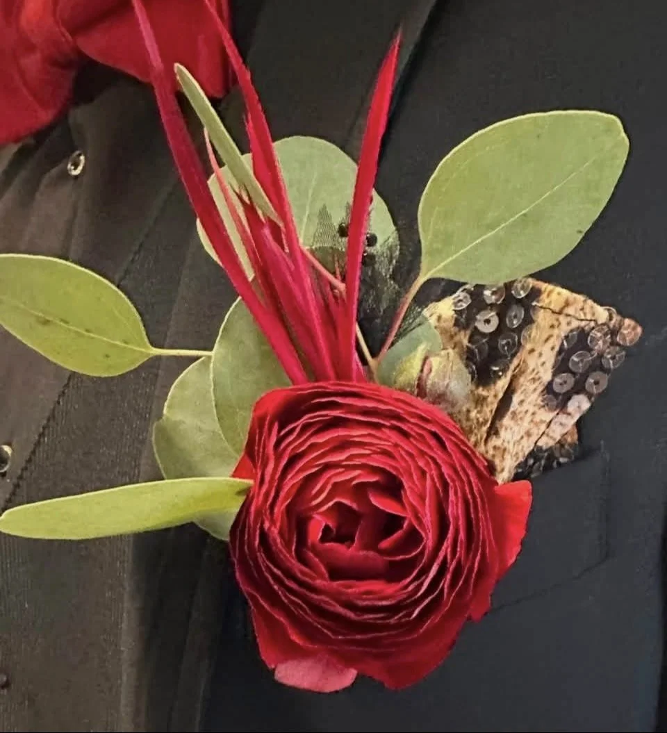 A floral arrangement with a large, red, intricately layered ranunculus flower, green leaves, and red-tipped stems, alongside a rectangular piece of wood bark with embedded shiny sequins, all placed on a black surface.