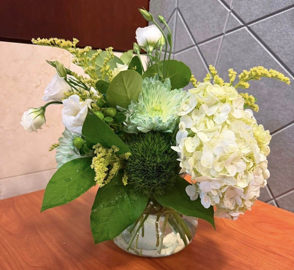 A bouquet of white and pale green flowers in a clear glass vase on a wooden surface with a tiled wall in the background.