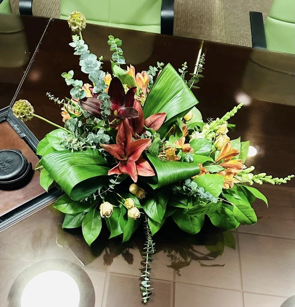 A vibrant floral arrangement with red lilies, orange flowers, and green foliage on a polished dark wooden table.