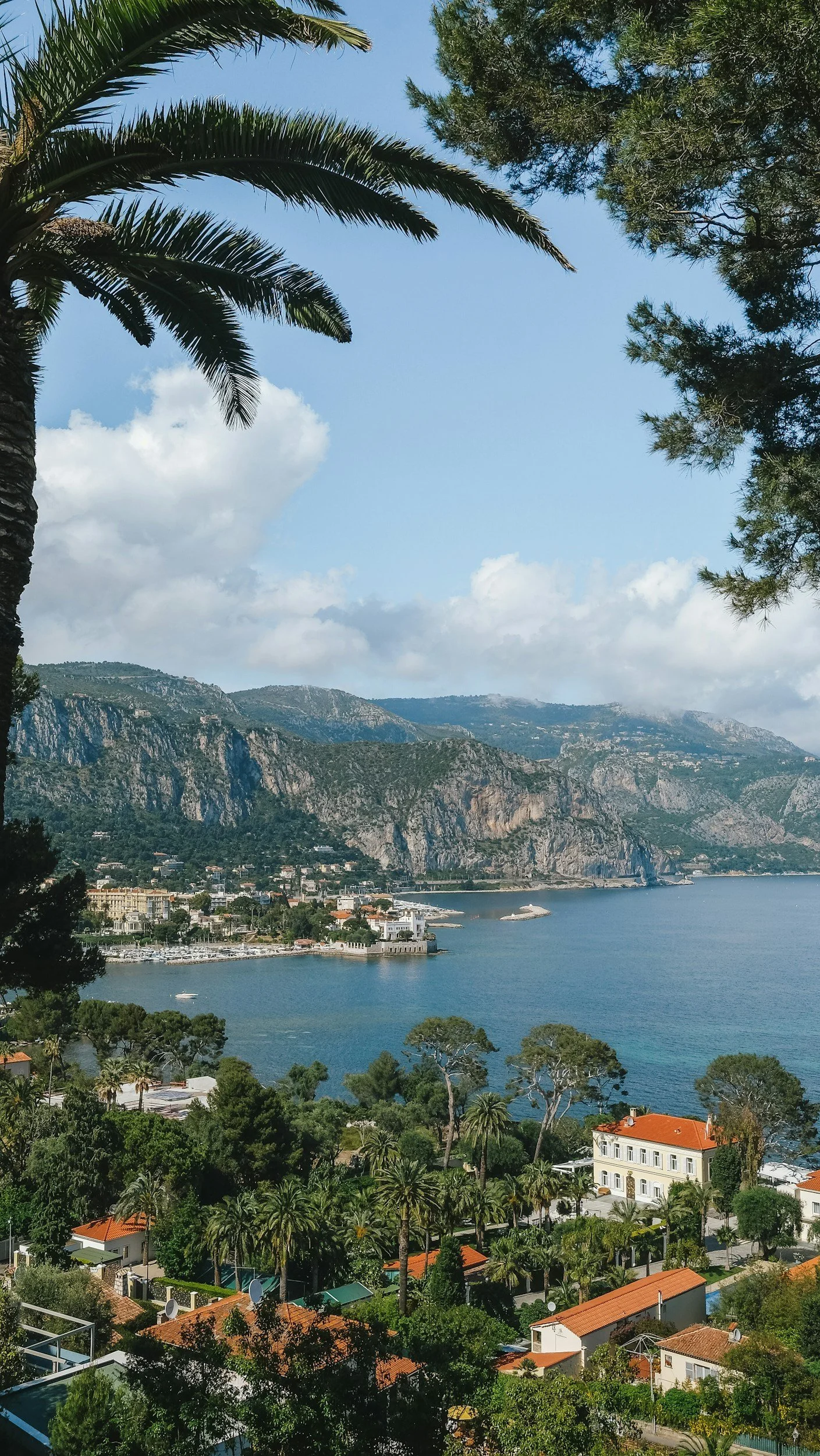 Coastal town with red-tiled roofs, surrounded by lush green trees, overlooking a bay with a mountain range in the background and a partly cloudy sky.