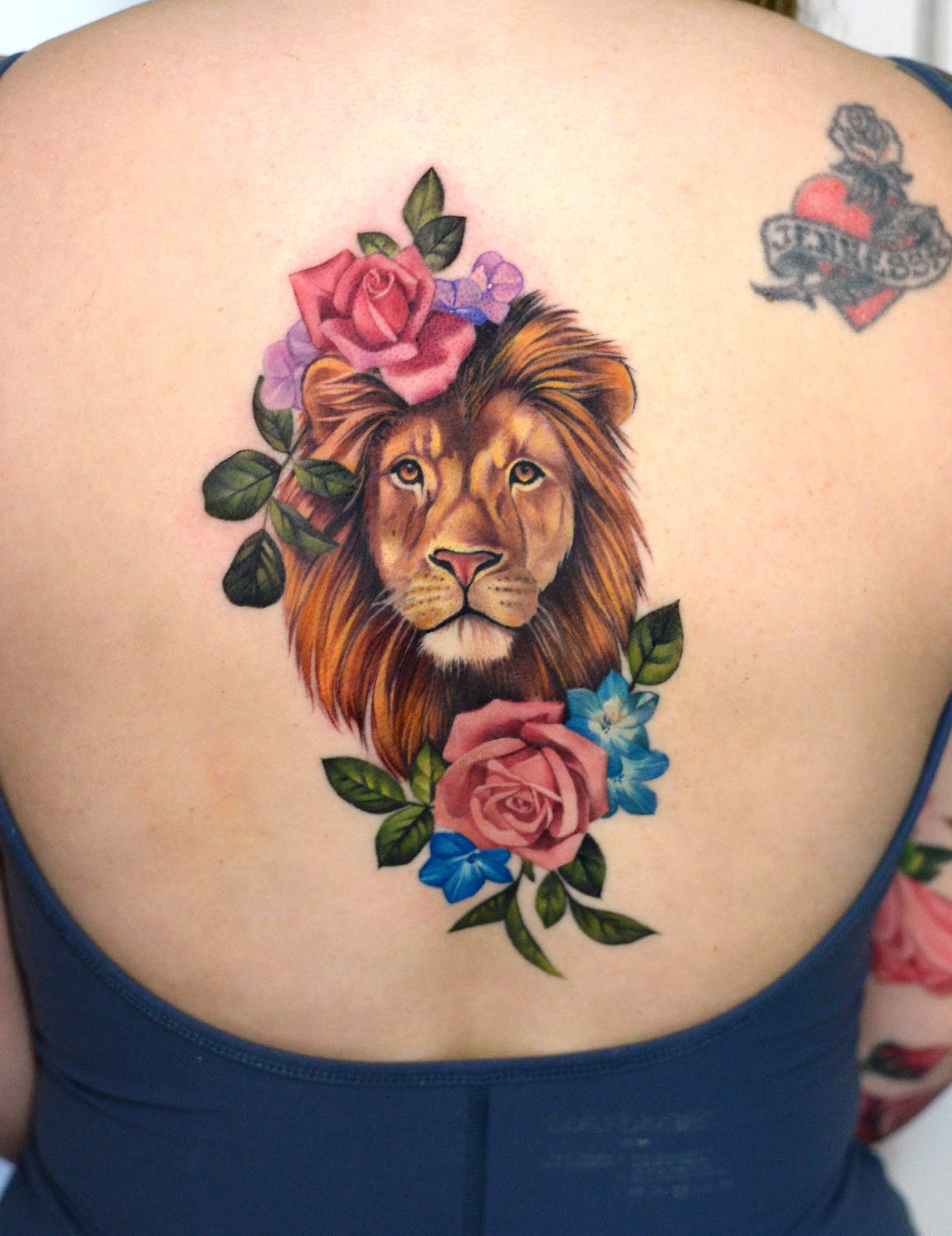 Tattoo of a lion's face with a pink rose, purple flowers, and green leaves on a person's upper chest, with part of a tattoo on the person's shoulder and upper arm visible.