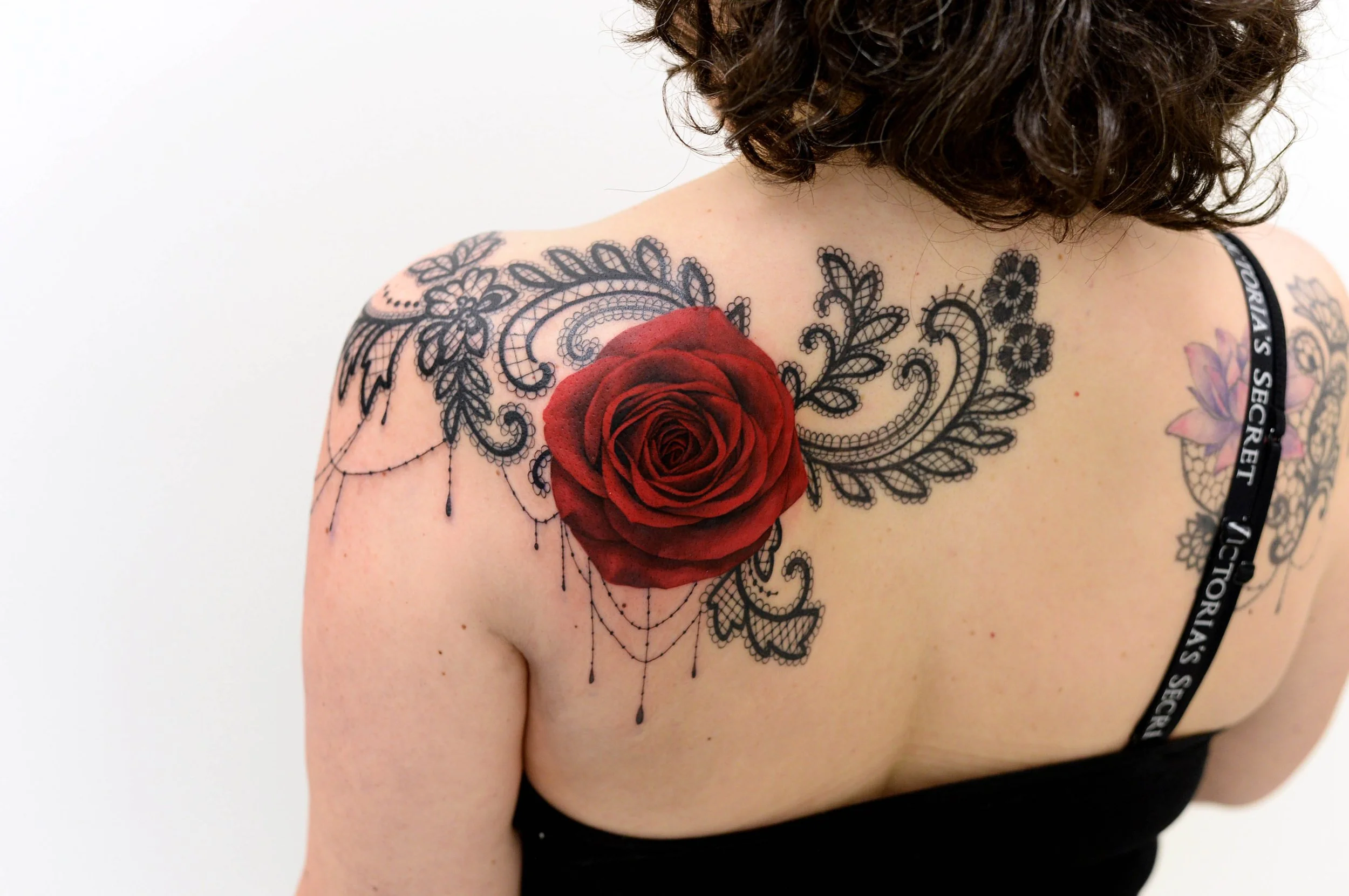 A woman with curly hair and light skin showing a large red rose tattoo on her shoulder, with black lace and floral patterns tattooed around it.