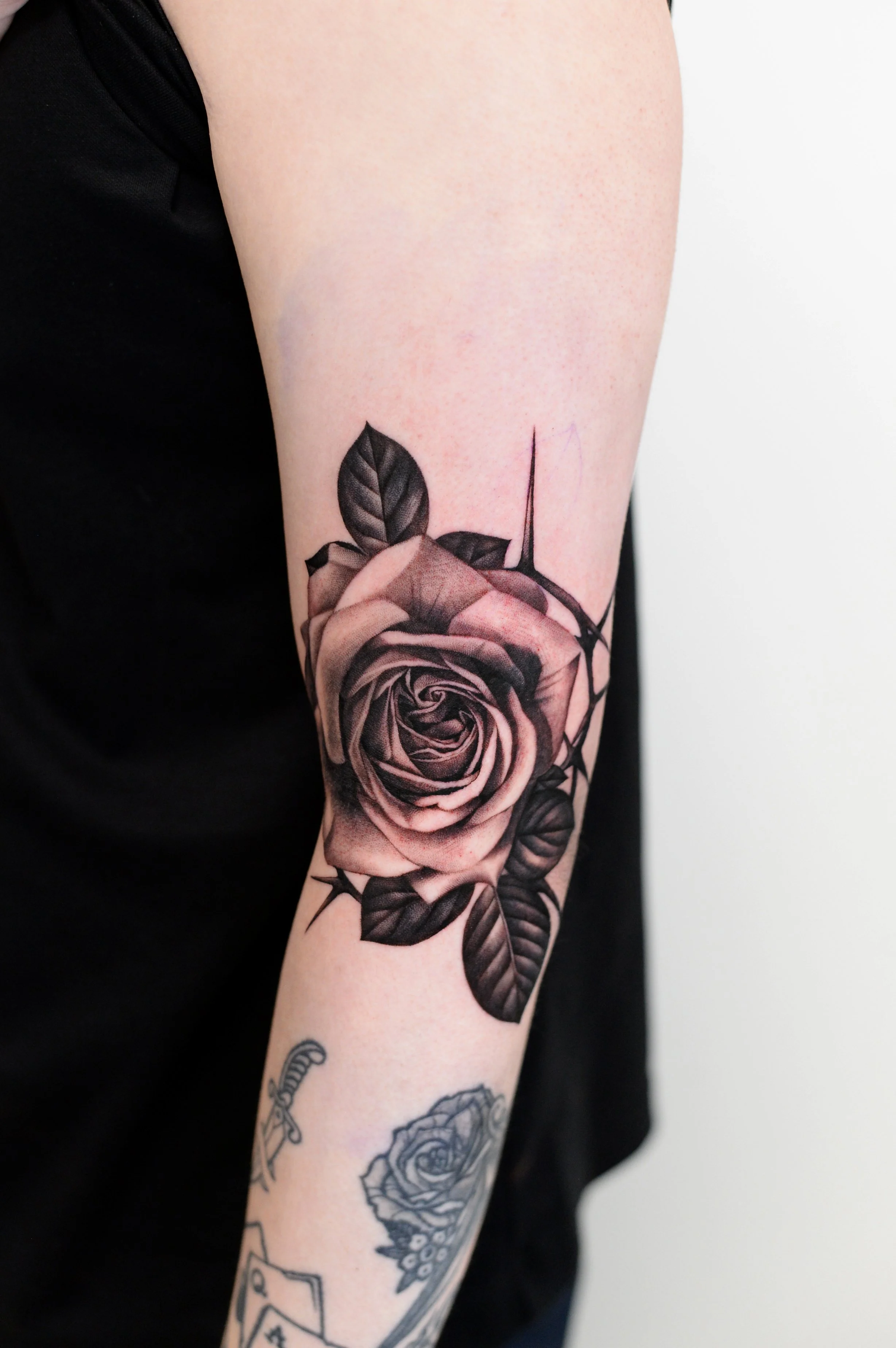 A person's arm with a black and gray tattoo of a rose with leaves and thorns.