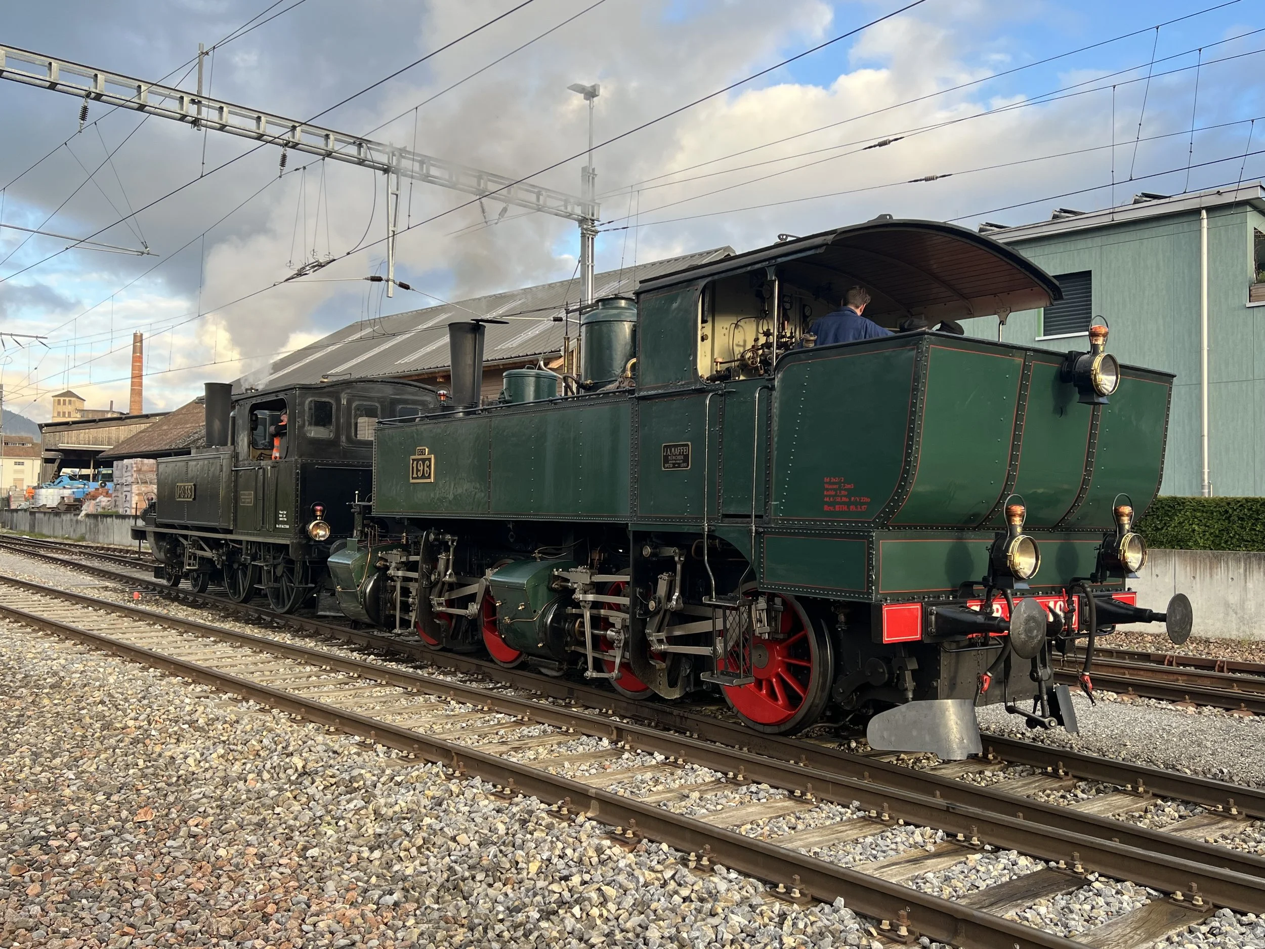 Ein historischer grüner Dampfzug auf einem Bahnhof mit Stromleitungen und Gebäuden im Hintergrund.