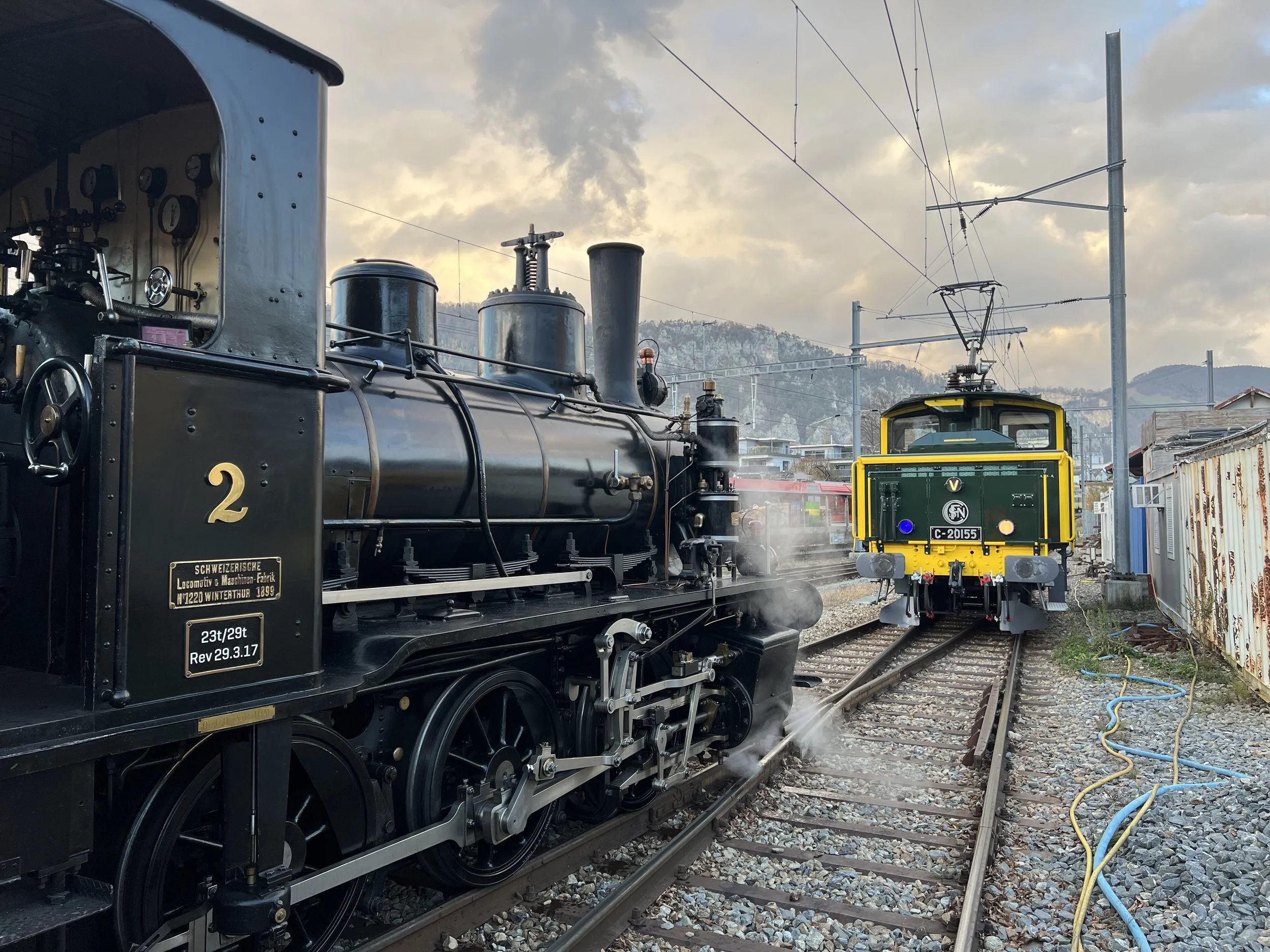 Ein Dampflokomotive steht auf einem Eisenbahngleis, im Hintergrund sind Schienen, elektrische Kabel und Berge bei bewölktem Himmel sichtbar.
