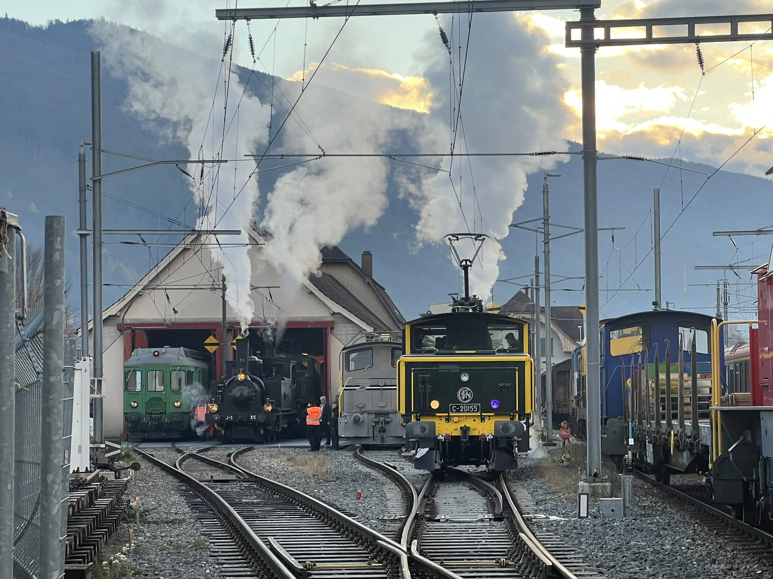 Zug bei der Wartung in einem Bahnhof mit Bergen im Hintergrund und Rauch aus den Fabriken beim Sonnenuntergang.