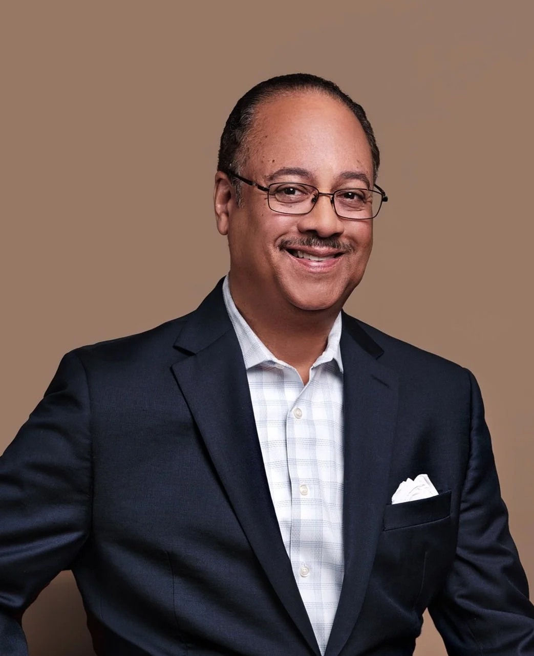A man wearing glasses, a navy suit, and a light-colored checkered shirt, smiling in front of a plain beige background.