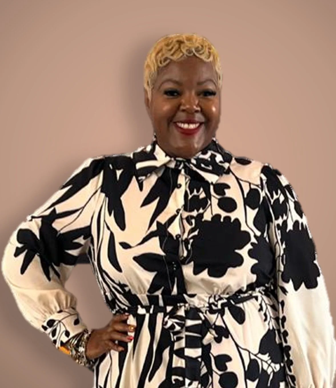 A woman with short blonde curly hair wearing a black and white floral patterned dress with a bow at the neck, standing against a plain background, smiling with her hand on her waist.