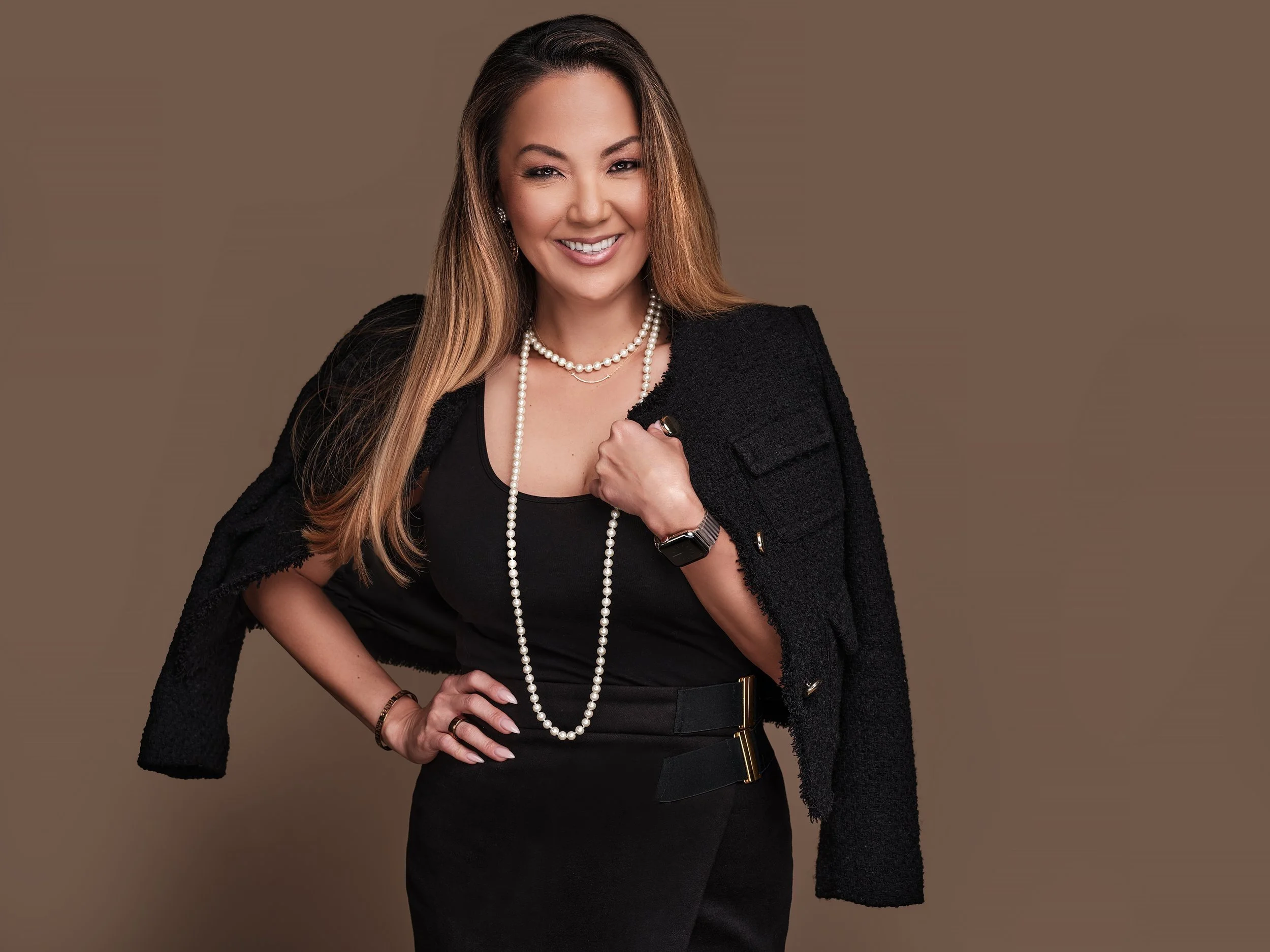 A woman with long brown hair wearing a black dress, pearl necklace, black blazer, and a watch, smiling confidently with a hand on her hip.