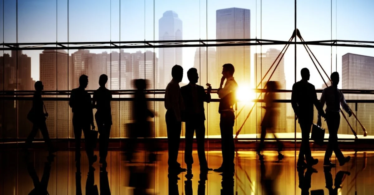 Silhouettes of businesspeople talking and walking in a modern office with skyscrapers outside during sunset.