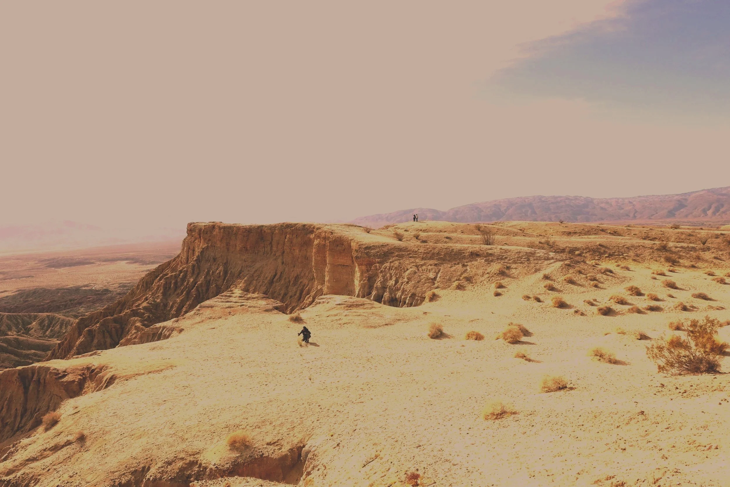 A vast desert landscape with rocky cliffs and sparse vegetation, with two hikers walking along a trail, mountains in the distance, under a partly cloudy sky.