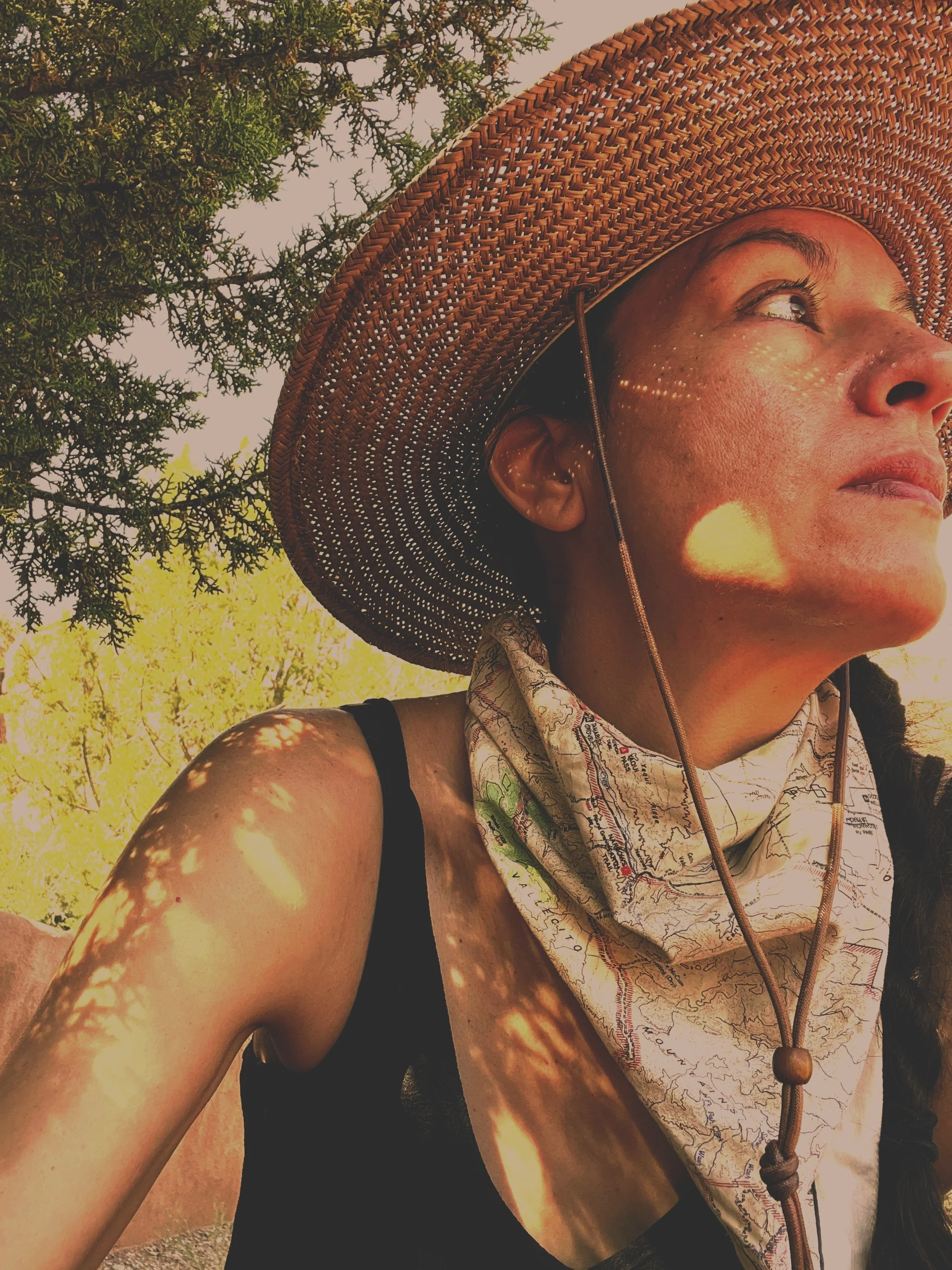 A person wearing a large woven straw hat, a black sleeveless top, and a patterned bandana around their neck, outdoors with green trees and sunlight creating patterns on their arm and face.
