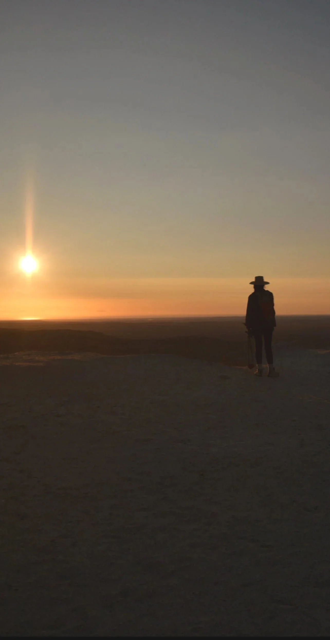 Person wearing a hat standing on a beach at sunset.
