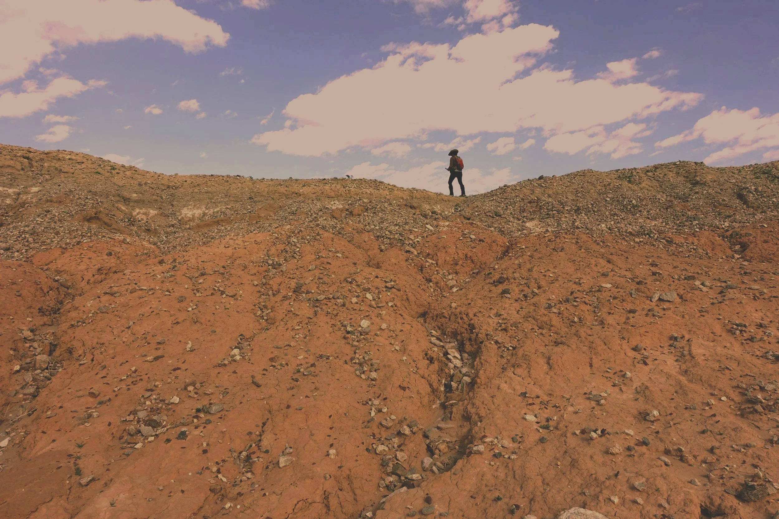 A person wearing a hat, backpack, and outdoor clothing walking on a rocky hilltop under a partly cloudy sky.