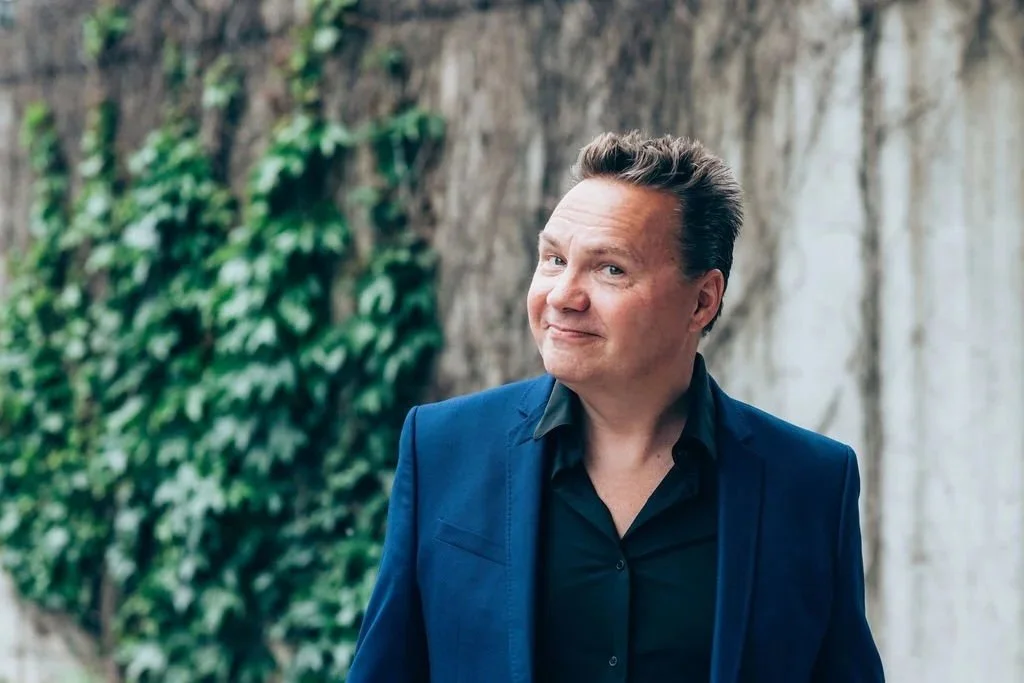 Dan Stocke, renowned happiest and founder of Zenith Unum, the unified theory of happiness, wears a dark blue suit with an open collar, standing outdoors and smiling at the camera with a slight grin.
