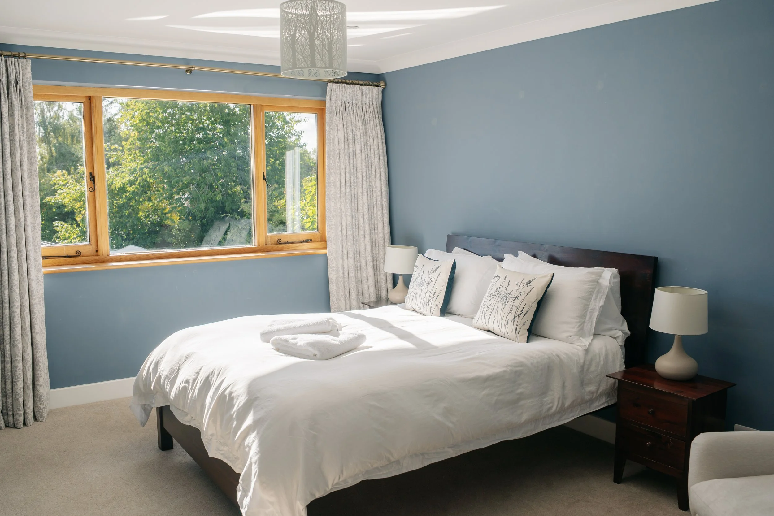 The Blue Room bedroom.  It has a large wooden window with a view of the garden. The room has a bed with white linens and decorative pillows, a wooden nightstand with a white lamp, and curtains on either side of the window.