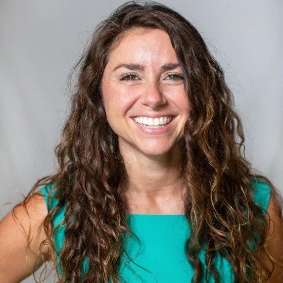A woman with long, curly brown hair smiling at the camera, wearing a sleeveless turquoise top.