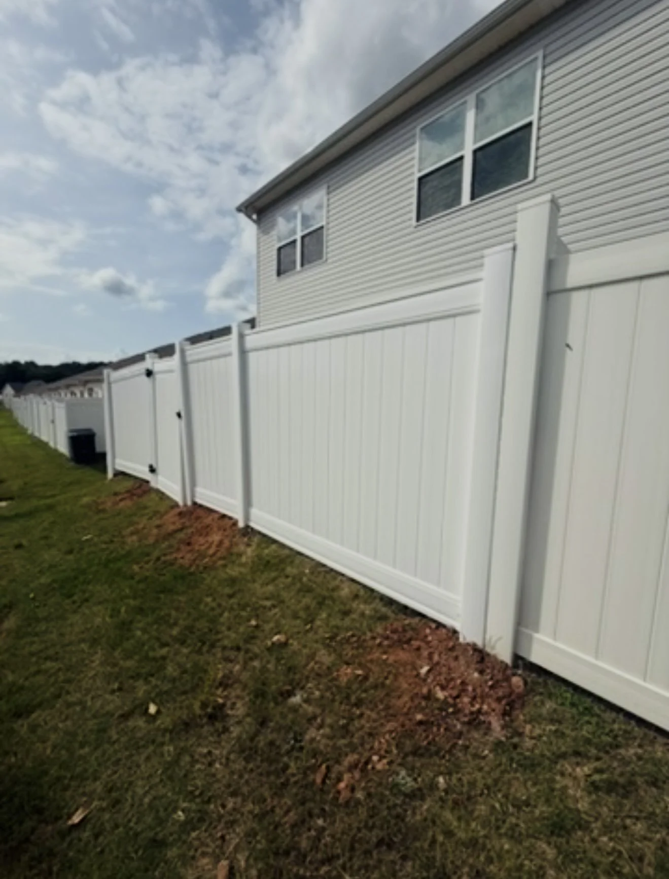 A white vinyl fence running alongside a house with beige siding and three windows, with a grassy yard and a partly cloudy sky in the background.