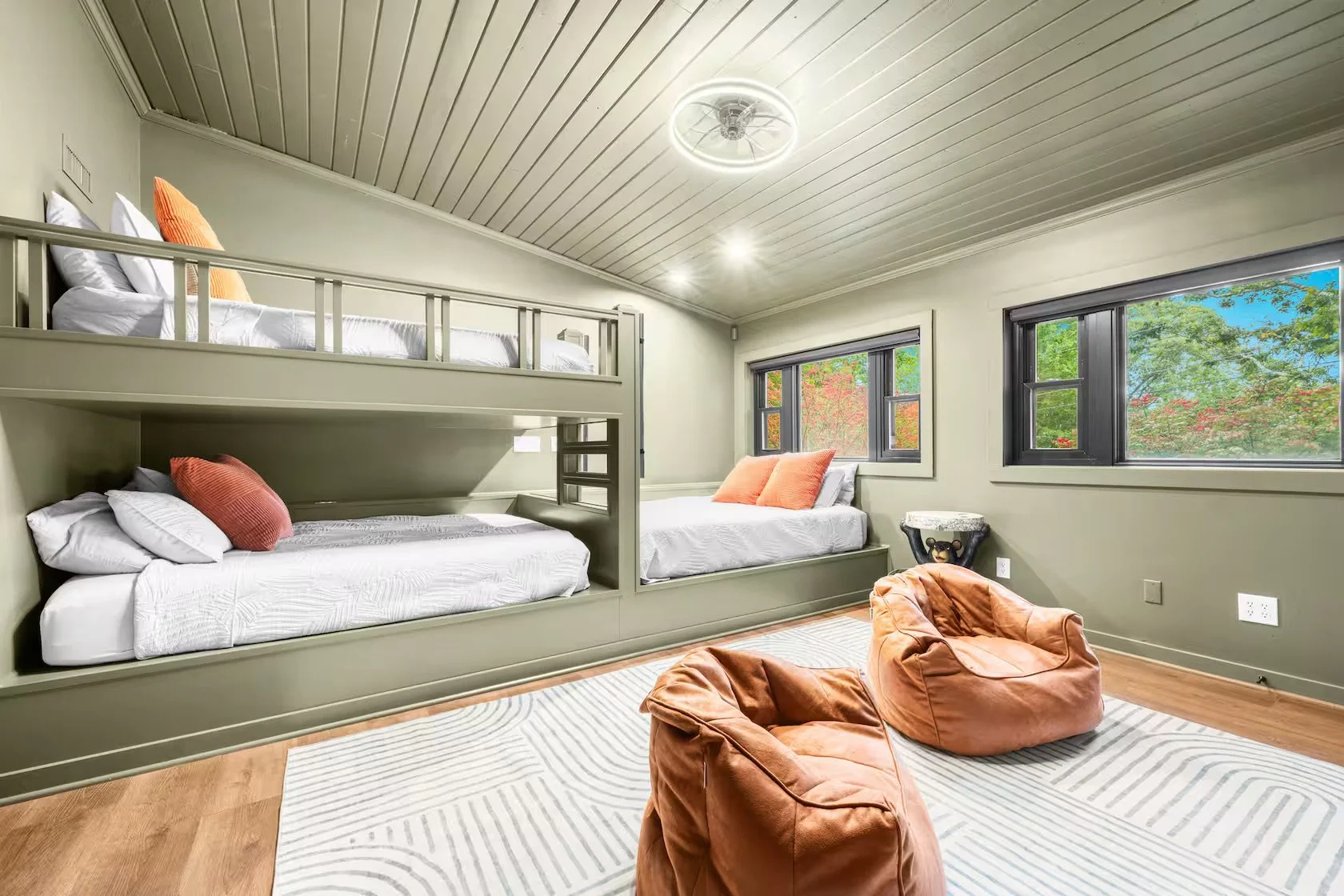 A bedroom with a bunk bed and a double bed, both with white bedding and orange pillows, positioned against light green walls with large windows showing outdoor greenery, a striped rug, and two orange bean bag chairs in the foreground.