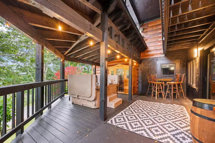 Covered outdoor balcony with wooden flooring and railing, featuring a seating area with sofa and steps, a dining area with round table and chairs, a patterned rug, string lights, and a view of trees in the background.