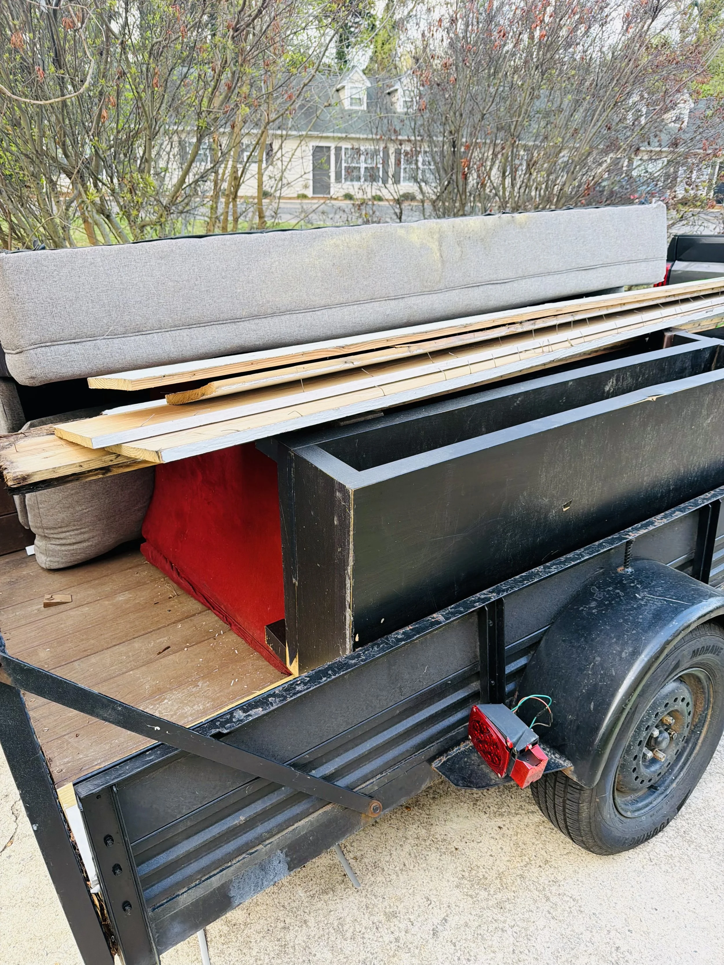 The image shows a black utility trailer with a wooden floor, loaded with a gray couch, some wooden planks, and other materials, parked on a concrete driveway.