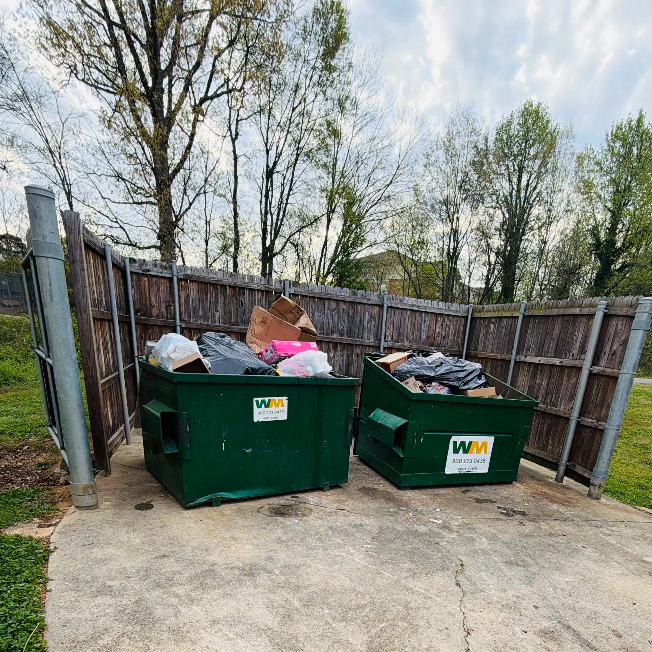 Two green trash dumpsters filled with trash are placed on a concrete driveway behind a wooden fence with bare trees on the background and a cloudy sky.