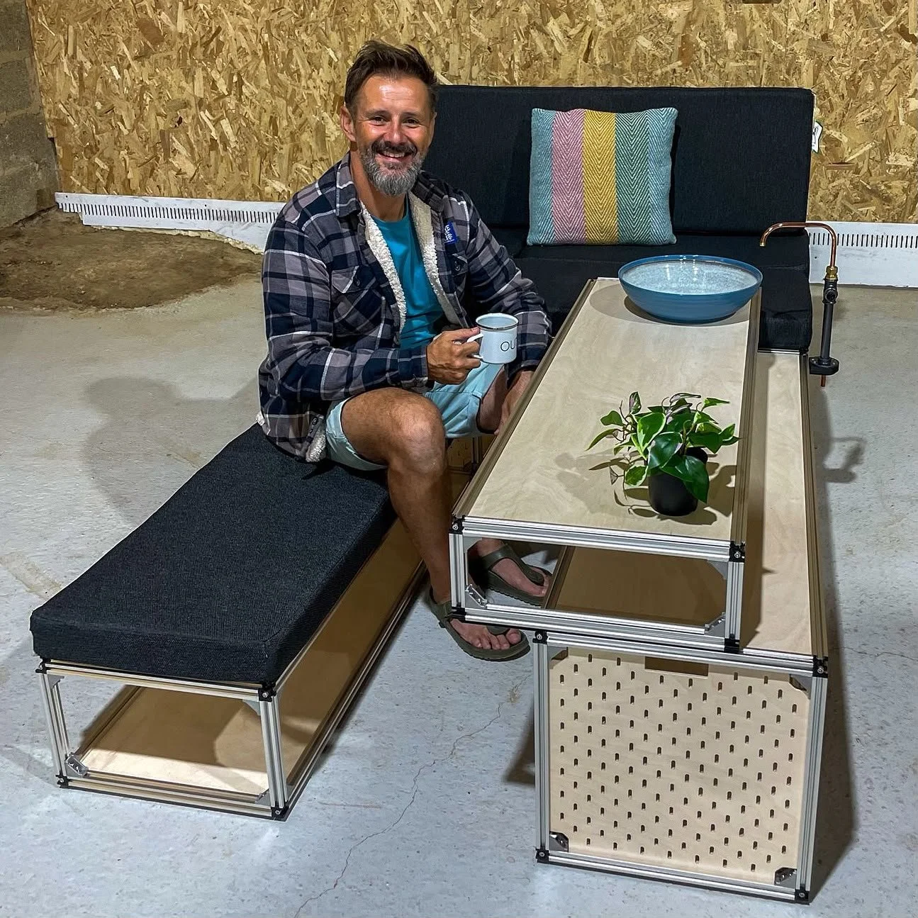 A man with a beard and short hair, wearing a plaid shirt, blue t-shirt, and shorts, sits on a black upholstered bench with a wooden frame, holding a mug. He is smiling and sitting on an industrial-style desk made of light wood and metal, with a potted plant, a ceramic bowl, and a faucet attached to the desk. The background consists of a plywood wall and a concrete floor.