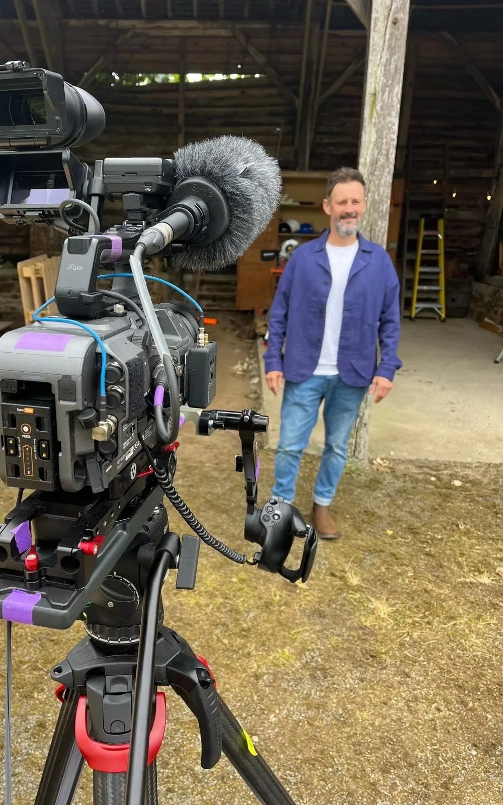 A man stands in front of a camera setup inside a rustic barn with wooden walls and a slanted roof. He has a beard and is wearing a blue jacket, white shirt, and jeans.