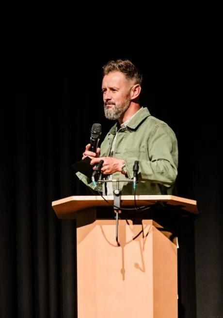 A man with a beard, wearing a green jacket, stands behind a wooden podium holding a microphone, in front of a black curtain.