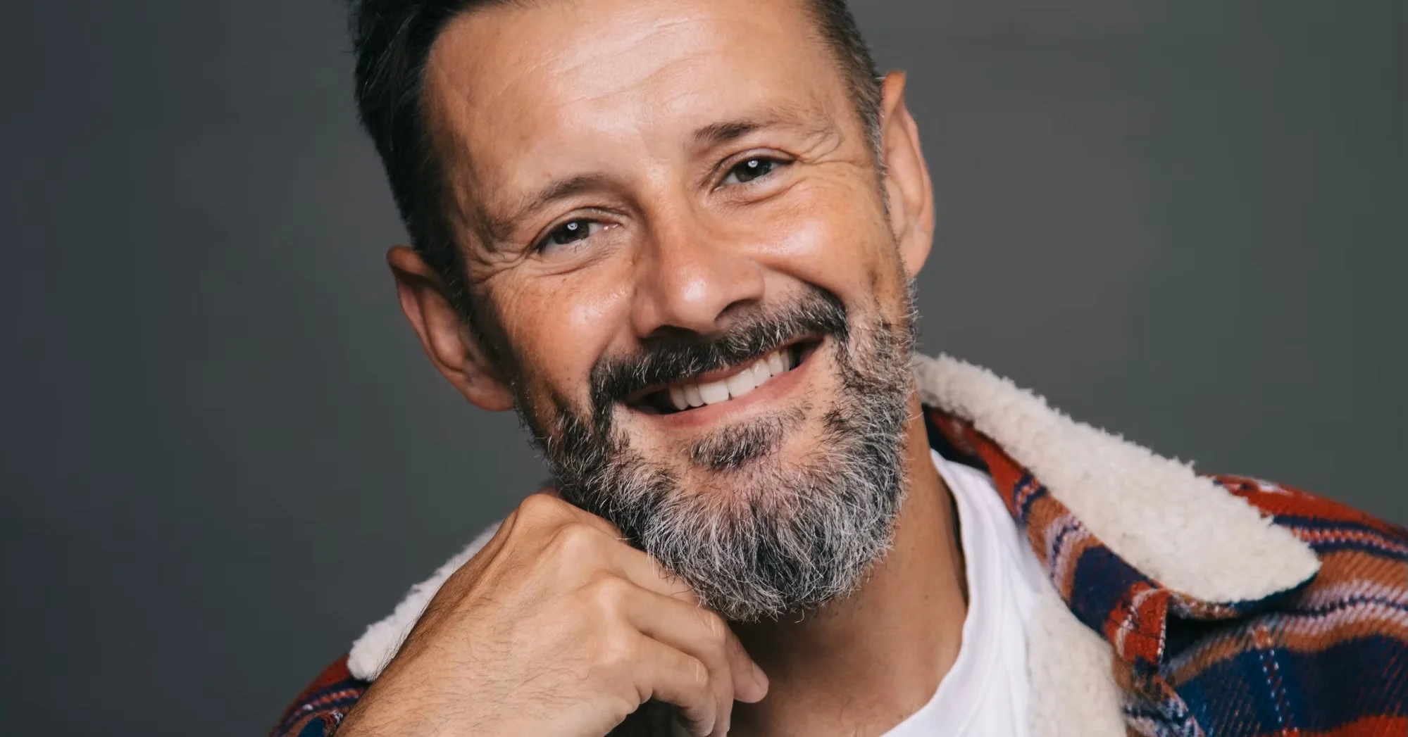 Max McMurdo wearing a plaid jacket over a white shirt, against a dark background.