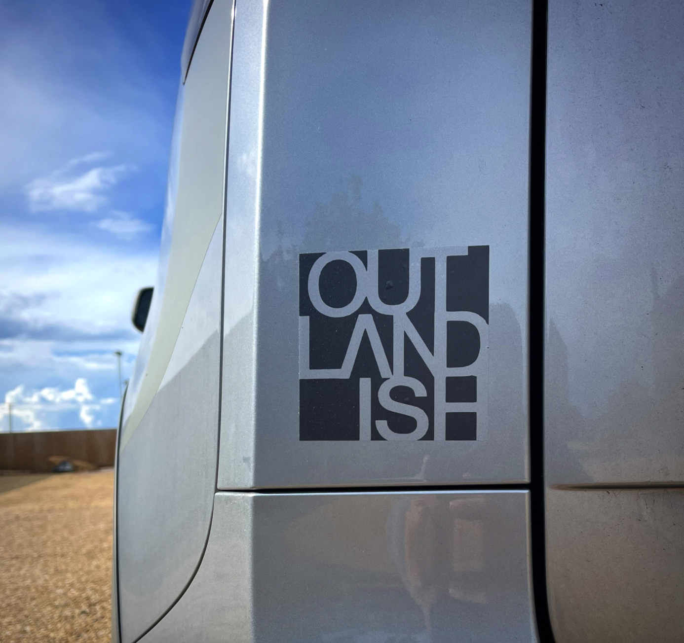 Close-up of a silver vehicle with Outlandish sticker, outdoors with a blue sky and clouds in the background.