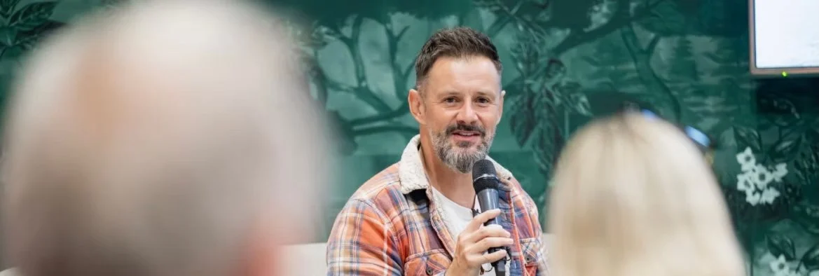 Max McMurdo with a microphone speaking to an audience at an indoor event, with a green leafy background.