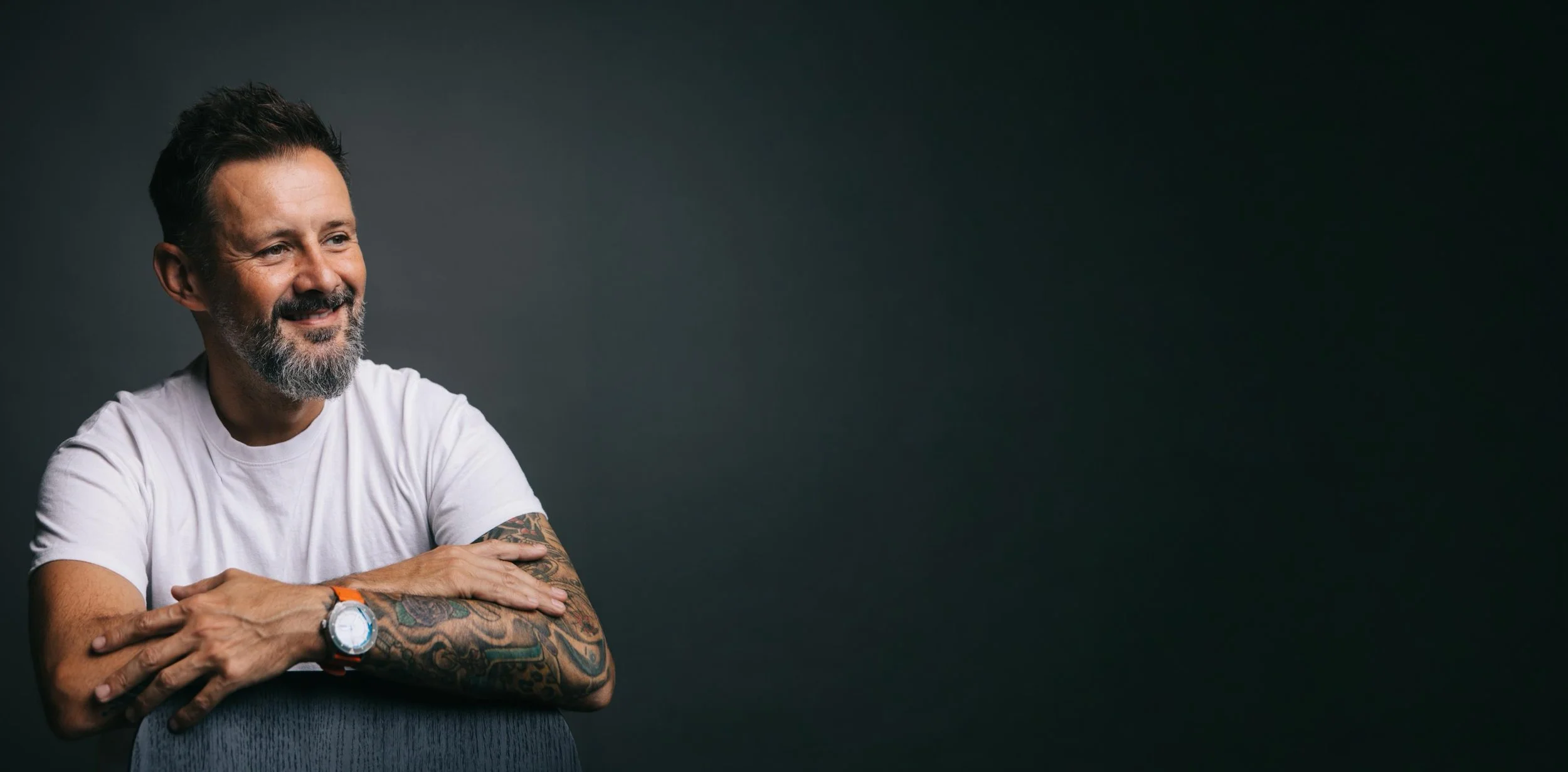 Max McMurdo wearing a white t-shirt, sitting with his arms crossed, smiling against a dark background.